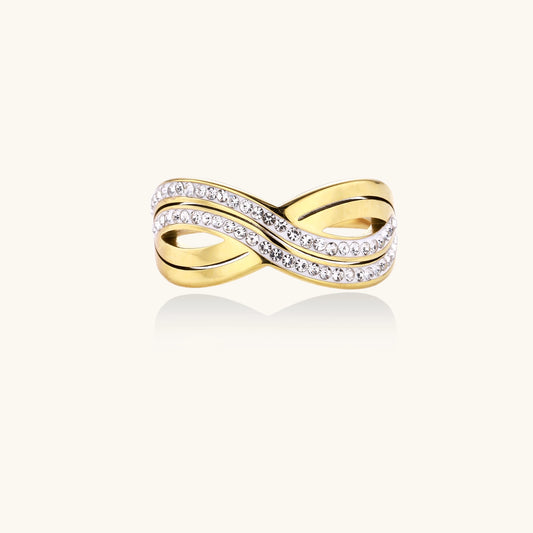Gold-plated stainless steel ring with crossed infinity bands adorned with sparkling rhinestones against cream background.