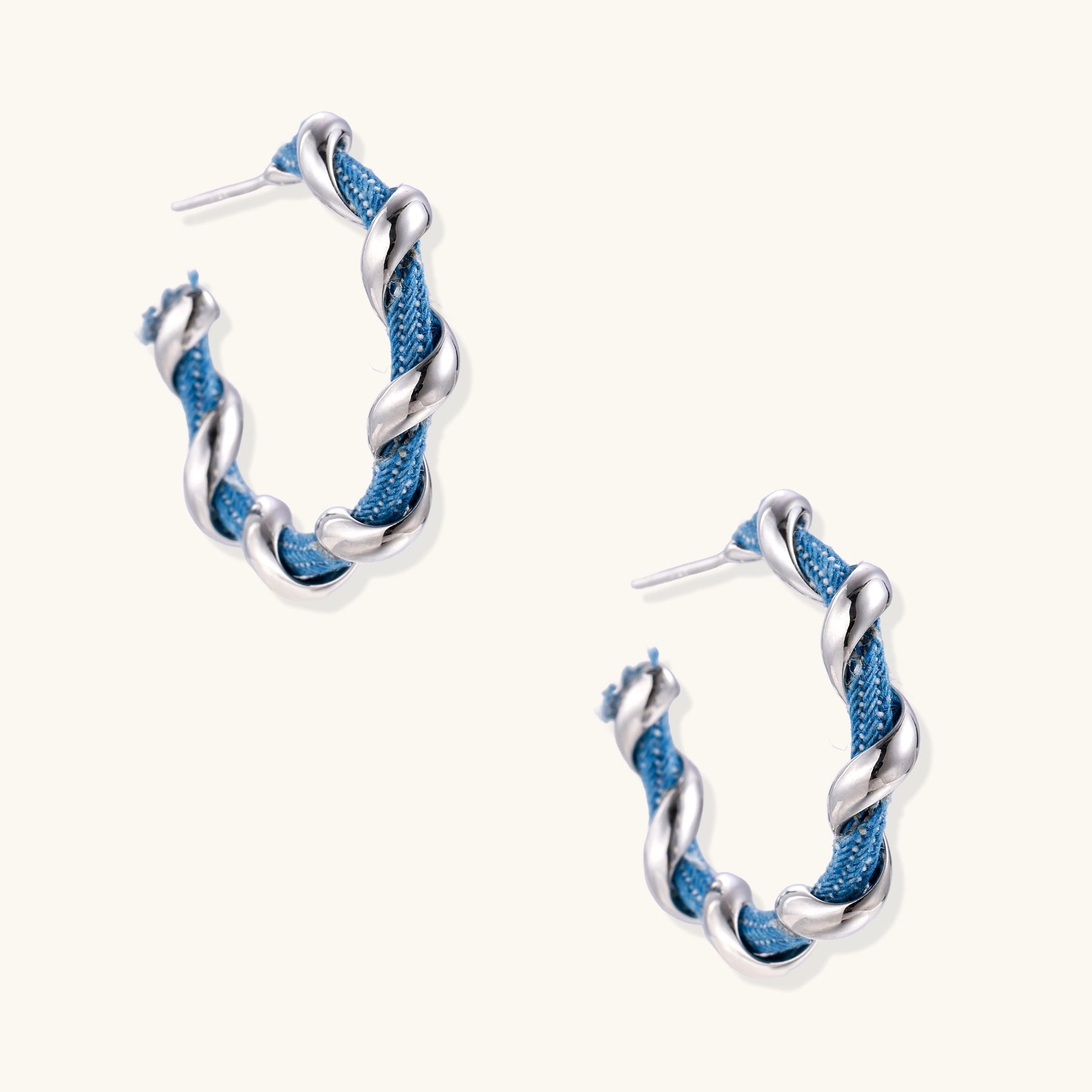 Twisted hoop earrings featuring blue denim fabric braided with gold-plated metal in a spiral design on white background.