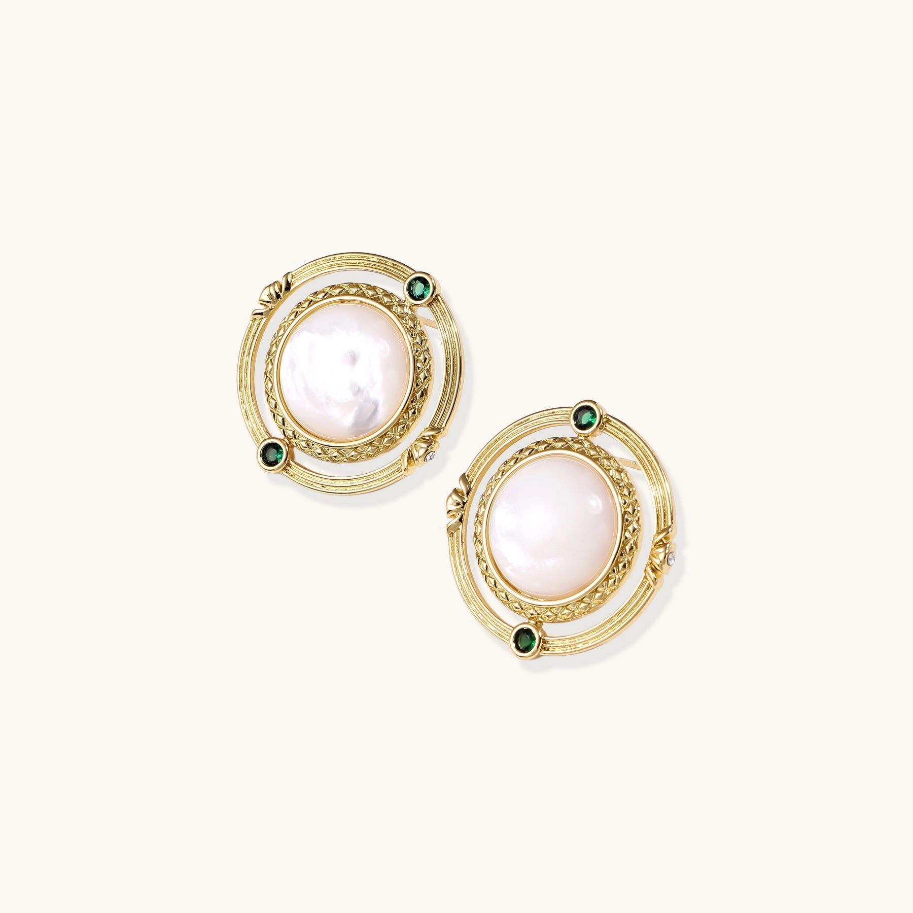 Close-up of gold-plated circular earrings with white mother-of-pearl centers and green zirconia accents on cream background.