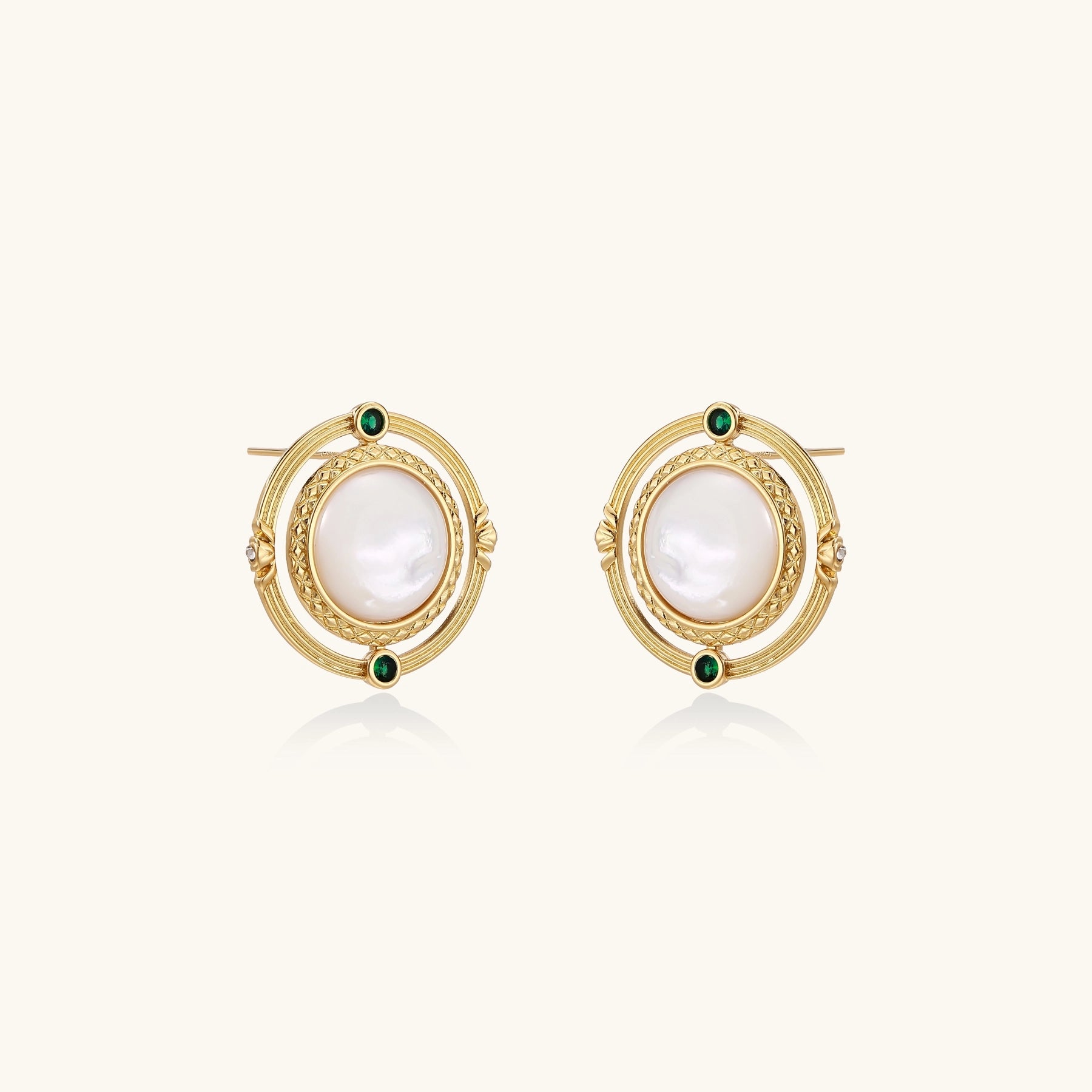 Close-up of gold-plated circular earrings with white mother-of-pearl centers and green zirconia accents on cream background.